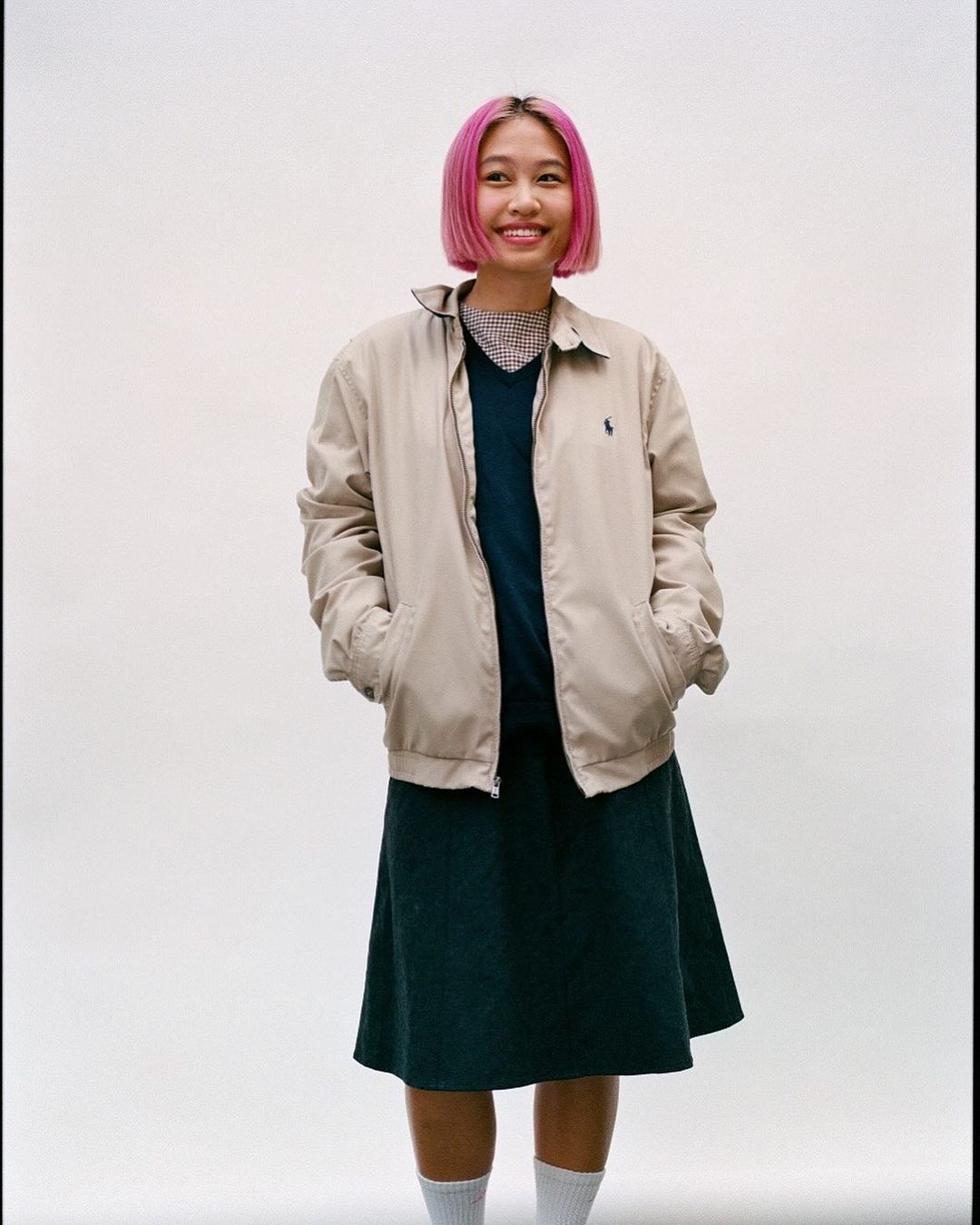 A full shot of a smiling woman with pink hair wearing a beige jacket, a dark skirt, and socks against a clean, neutral backdrop.