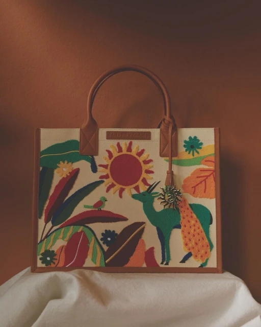 Stylish tote bag featuring nature-inspired embroidered artwork: a sun, a peacock, a gazelle, tropical plants, on an off-white background.