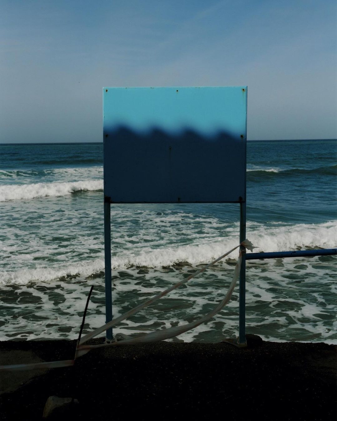 A rectangular sign painted with blue and dark blue wave patterns stands outdoors against a real ocean and clear sky background.