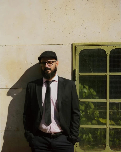 Portrait of a bearded man in a suit and cap standing against a textured wall next to a window with green foliage visible inside.