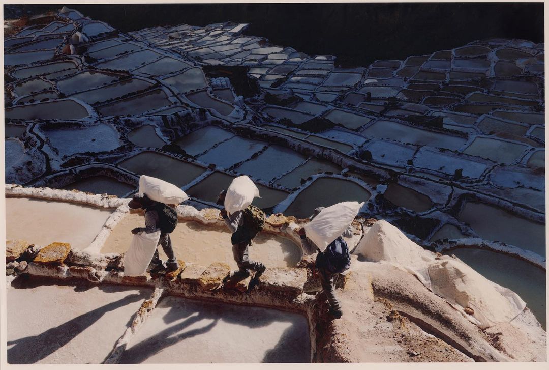 Three people carry sacks across salt ponds in Peru, captured in a sunny, medium outdoor shot with a clear view of the landscape.