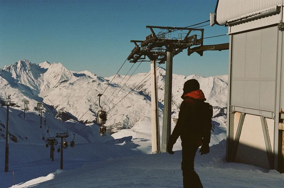 Person in black jacket stands near a ski lift on snow-covered mountain against a clear blue sky, with chairlifts and a station.