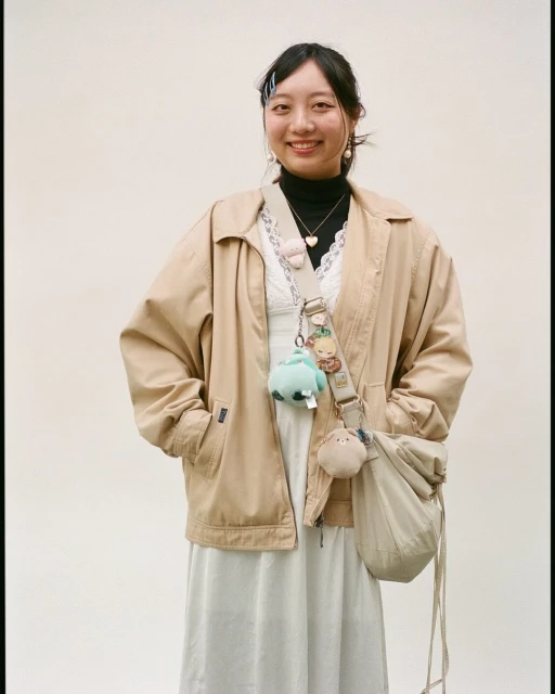 Portrait of a smiling Asian woman in a light-brown jacket and light-colored dress with accessories, standing against a plain background.