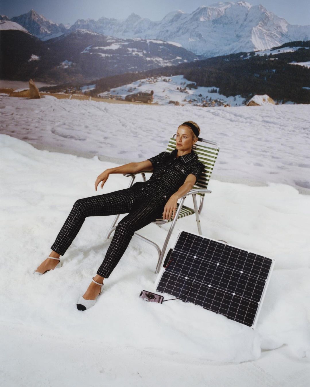 Woman in a black outfit reclines on a beach chair next to a solar panel and mobile phone, snow on the ground and mountains in the background.