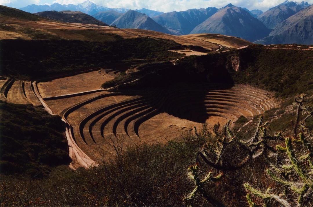 Terraced circular depressions at Moray archaeological site, Peru, against a backdrop of mountains and blue sky with clouds.