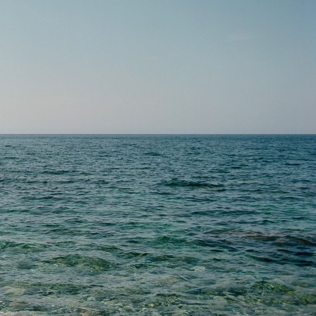 Overlooking calm blue ocean waters stretching to meet a pale blue sky at the horizon, seascape with serene and minimal aesthetic.