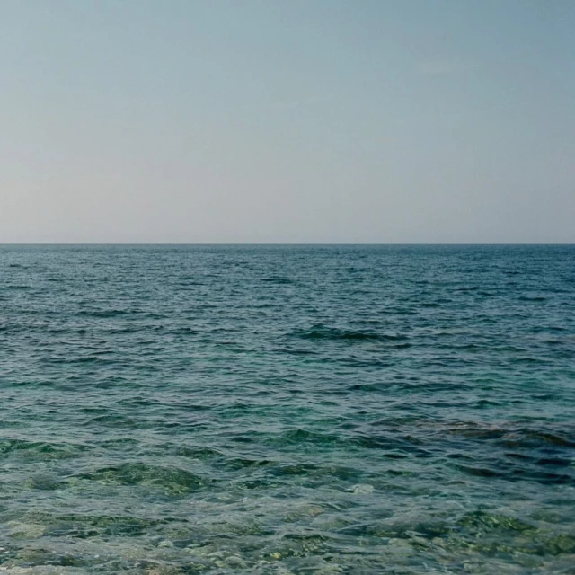 Overlooking calm blue ocean waters stretching to meet a pale blue sky at the horizon, seascape with serene and minimal aesthetic.