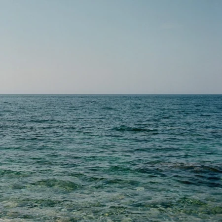 Overlooking calm blue ocean waters stretching to meet a pale blue sky at the horizon, seascape with serene and minimal aesthetic.