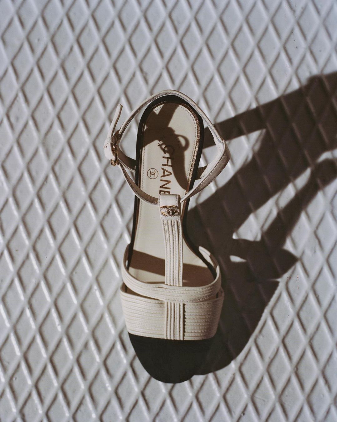 Elevated view of a cream-colored Chanel heeled sandal with an ankle strap on a white diamond-patterned surface casting a shadow.