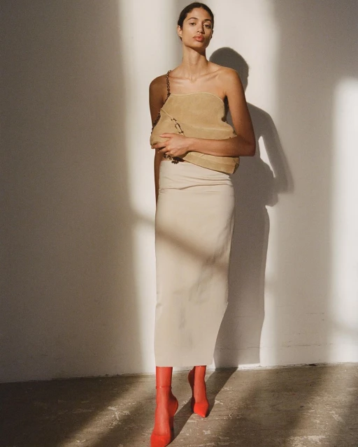Indoor shot of a stylish adult woman wearing a beige dress, red shoes, and carrying a beige bag with chain strap, posing in front of a white wall.