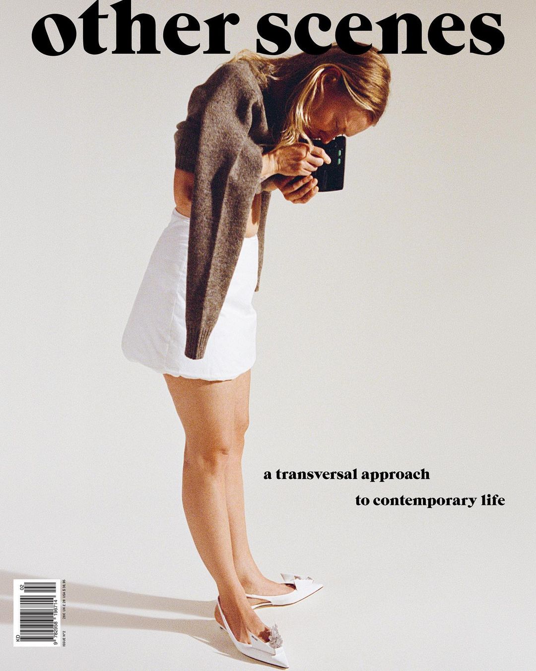 Blonde woman in white skirt holding device in white studio, magazine cover with 'other scenes' headline, modern fashion and lifestyle.