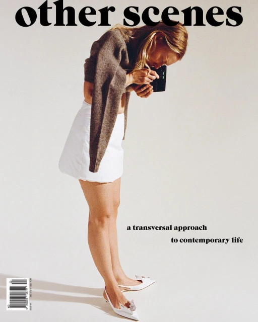 Blonde woman in white skirt holding device in white studio, magazine cover with 'other scenes' headline, modern fashion and lifestyle.