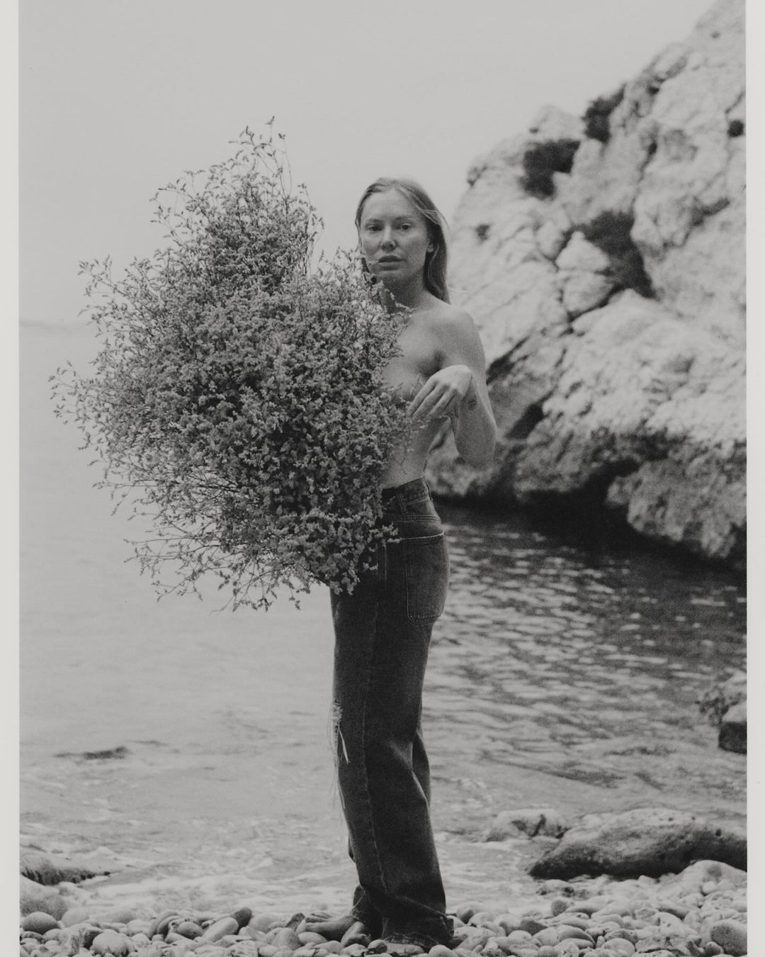 Black and white shot of a woman holding a large bunch of foliage on a pebble beach with the ocean and rocks in the background.