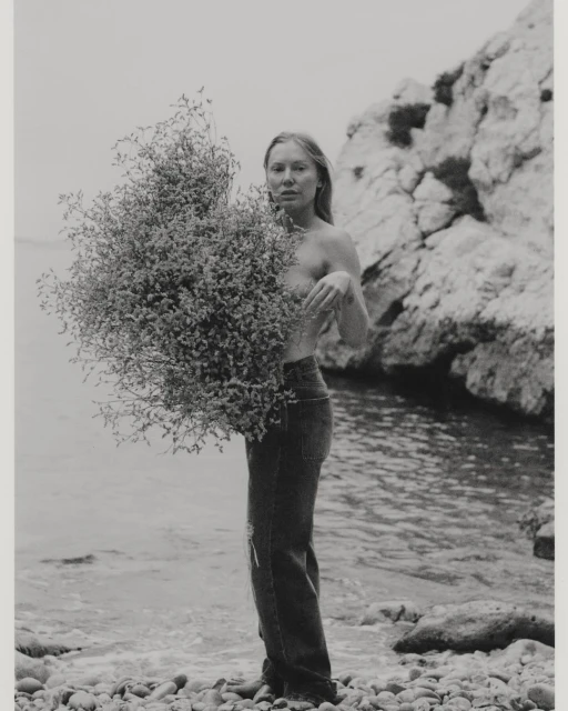 Black and white shot of a woman holding a large bunch of foliage on a pebble beach with the ocean and rocks in the background.
