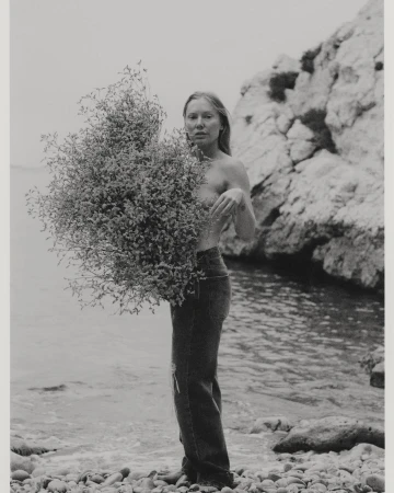 Black and white shot of a woman holding a large bunch of foliage on a pebble beach with the ocean and rocks in the background.
