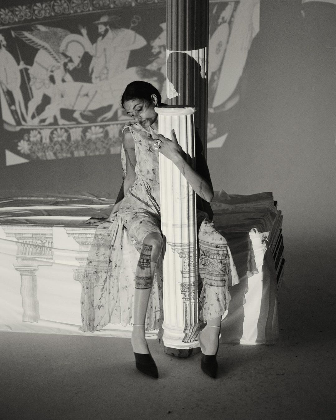 A woman in a floral dress is sitting and leaning against a column, an ancient Greek mural projected onto the background wall.