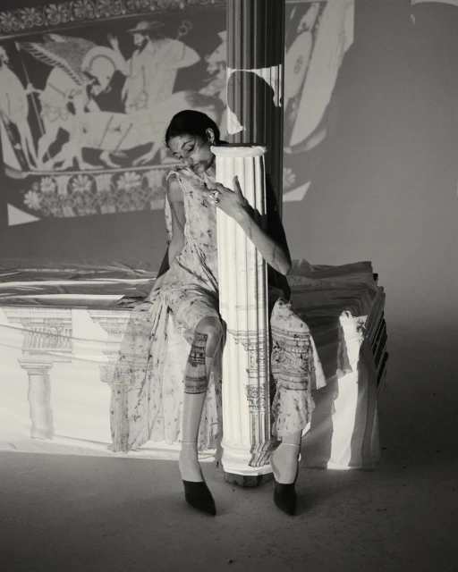 A woman in a floral dress is sitting and leaning against a column, an ancient Greek mural projected onto the background wall.