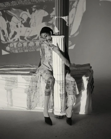 A woman in a floral dress is sitting and leaning against a column, an ancient Greek mural projected onto the background wall.