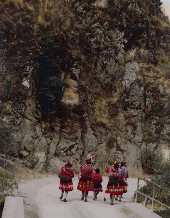 A group of Indigenous women and a child walk along a dirt road in a mountain landscape, wearing traditional clothing with red and black patterns.
