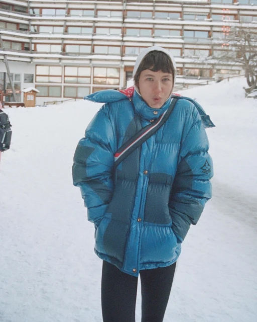 Woman in a blue puffer jacket and white headscarf stands in snow, posing outdoors near a multi-story building on a winter day.