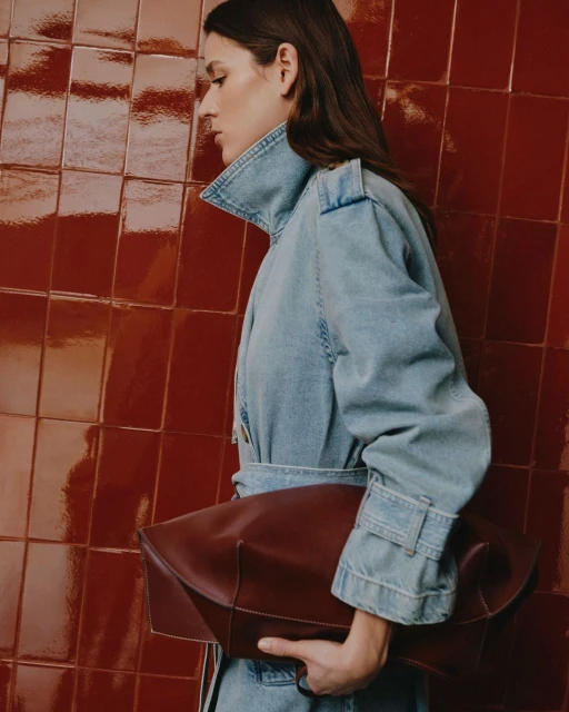 Adult female in a denim jacket holds a large leather bag against a textured tile wall in a studio setting. Fashion editorial.