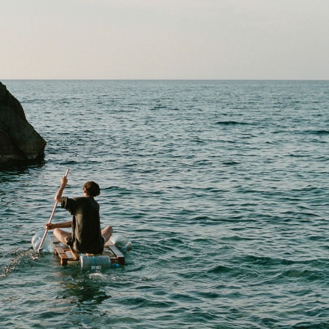 Person paddles makeshift raft with a long pole in the ocean, with a rocky land formation visible in the background against a light sky.