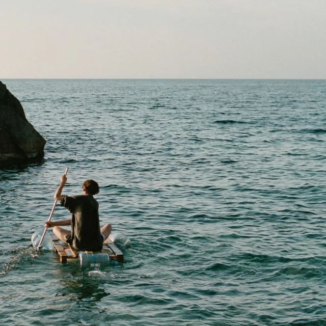 Person paddles makeshift raft with a long pole in the ocean, with a rocky land formation visible in the background against a light sky.