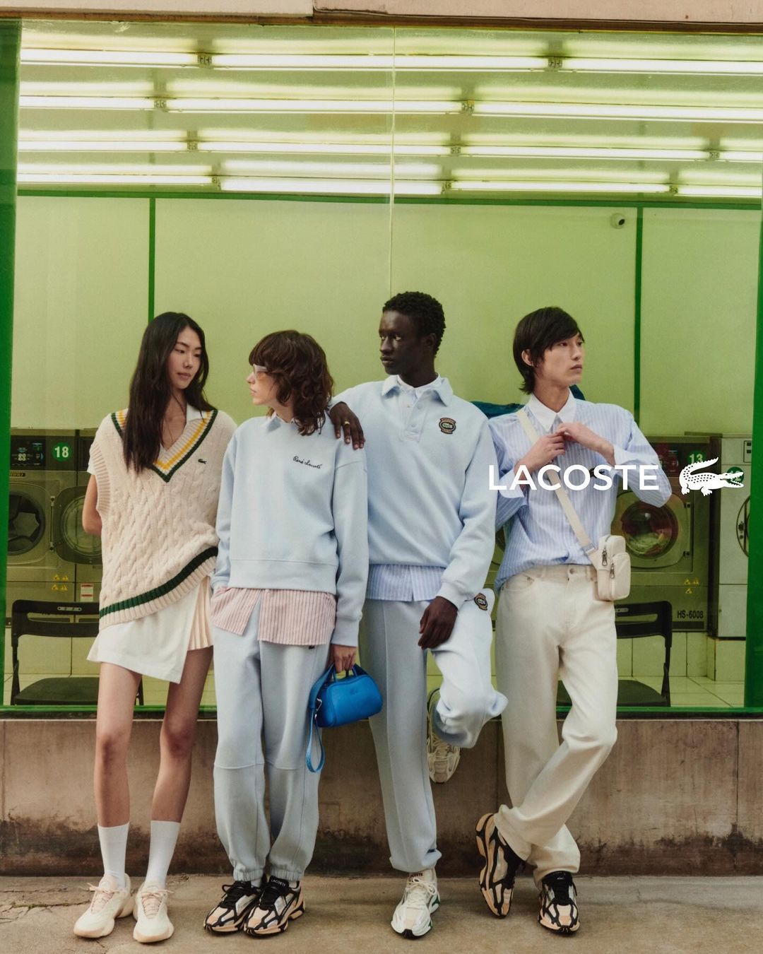 Four diverse young adults in Lacoste apparel pose in front of a laundromat, showcasing casual wear with a blend of light colors and urban style.