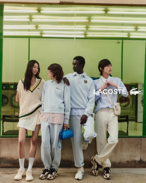 Four diverse young adults in Lacoste apparel pose in front of a laundromat, showcasing casual wear with a blend of light colors and urban style.