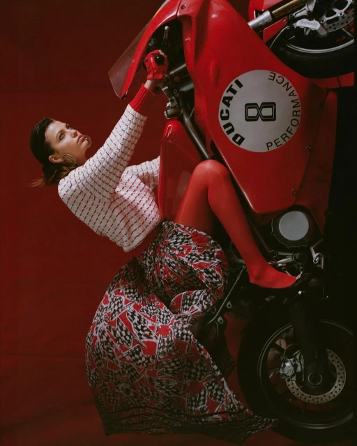 Glamour shot of woman in skirt straddling red Ducati motorcycle in studio with red background. Automotive fashion, motorcycle lifestyle.
