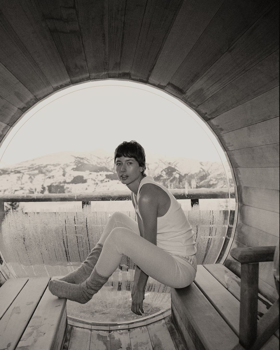 Monochrome photograph of a young adult sitting on a wooden bench inside of a sauna, with a snow-covered landscape in the background.
