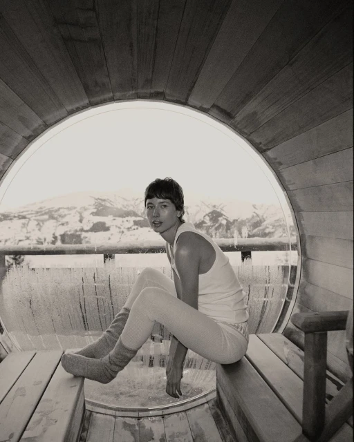 Monochrome photograph of a young adult sitting on a wooden bench inside of a sauna, with a snow-covered landscape in the background.