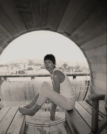 Monochrome photograph of a young adult sitting on a wooden bench inside of a sauna, with a snow-covered landscape in the background.