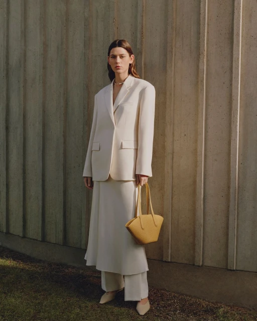 Woman in an off-white suit poses in front of a textured wall, carrying a yellow-tan woven handbag, in a medium shot with outdoor light.