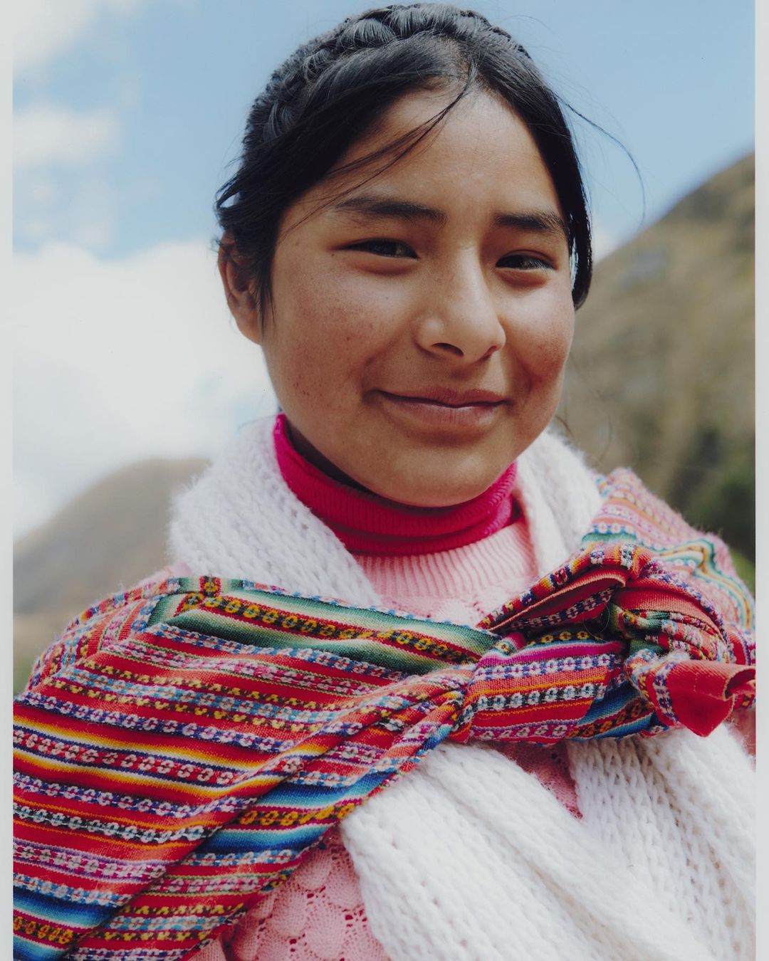 Portrait of a young woman with dark hair in a braided style, wearing a colorful woven scarf and pink sweater, smiles at the camera outdoors.