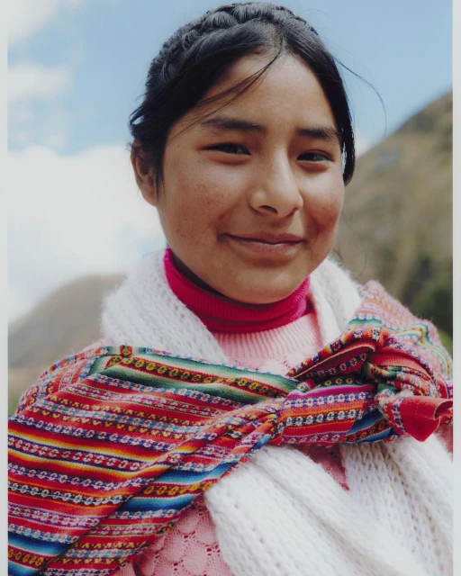 Portrait of a young woman with dark hair in a braided style, wearing a colorful woven scarf and pink sweater, smiles at the camera outdoors.