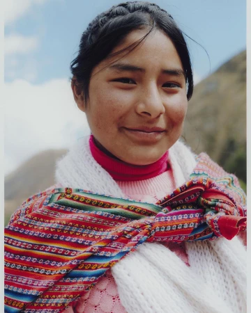 Portrait of a young woman with dark hair in a braided style, wearing a colorful woven scarf and pink sweater, smiles at the camera outdoors.