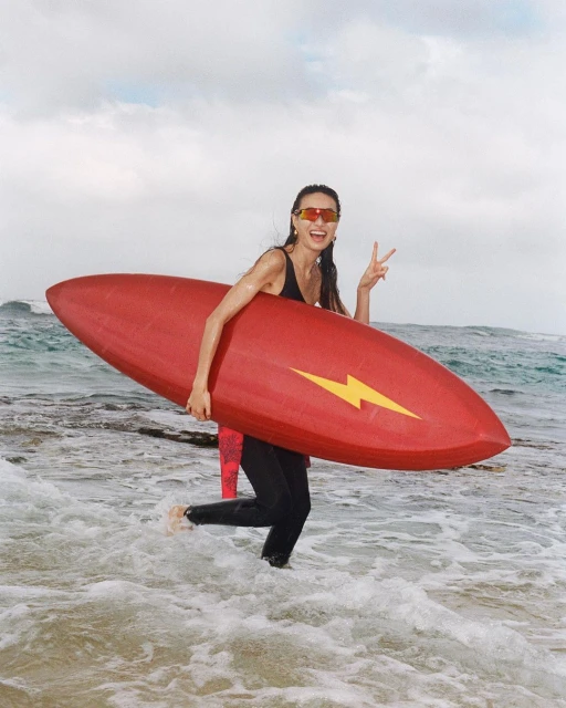 Smiling woman in ocean with a red surfboard featuring yellow lightning bolt; wearing sunglasses and a black wetsuit.