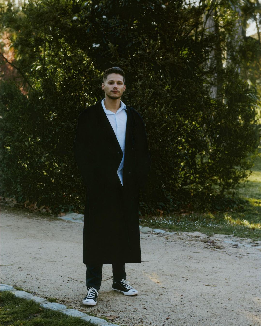 Man wearing a long black coat and sneakers standing on a sandy path before green bushes