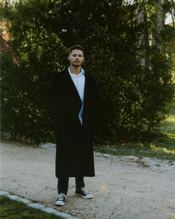 Man wearing a long black coat and sneakers standing on a sandy path before green bushes