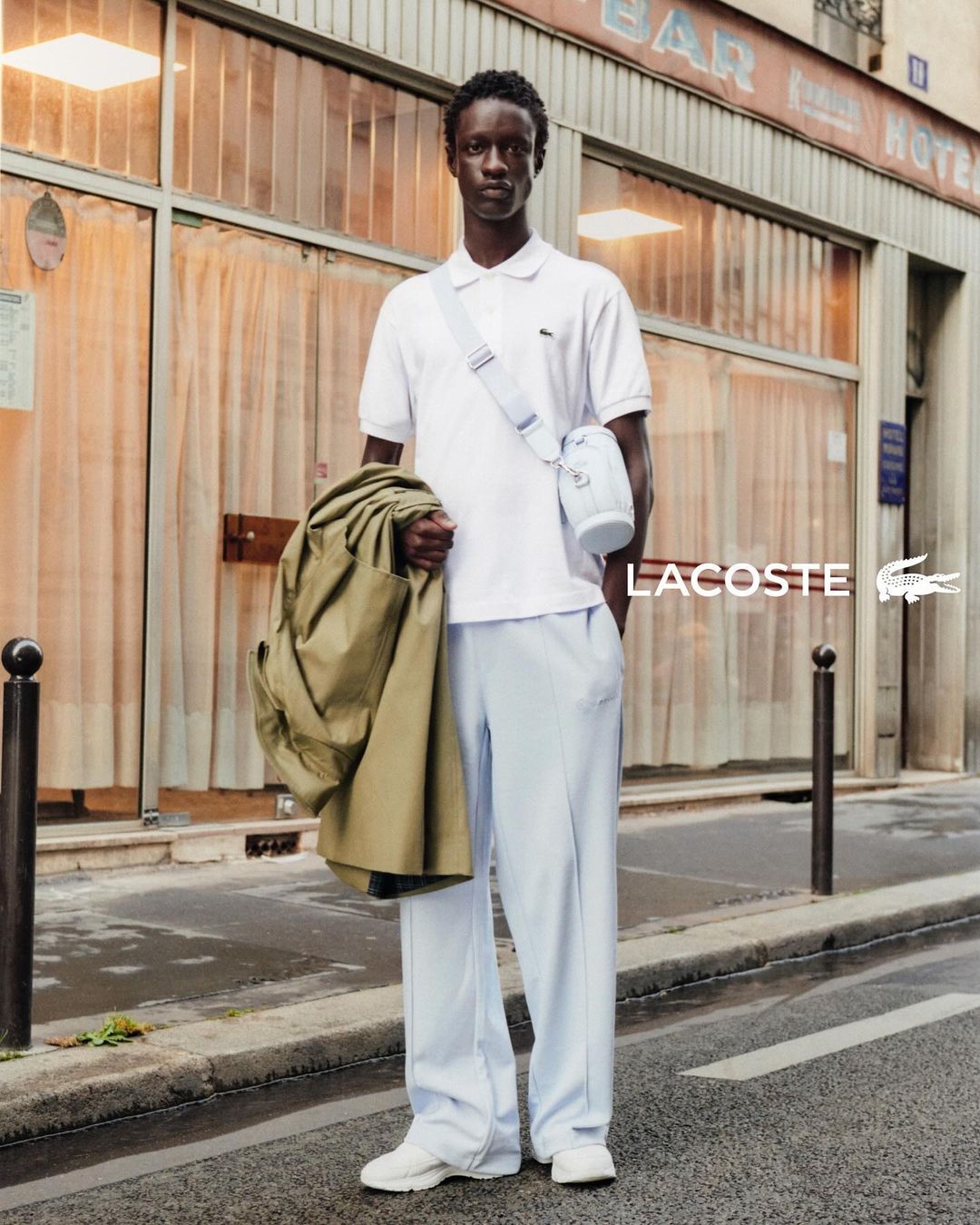 Adult male model in polo shirt and trousers posing in front of European urban storefront with Lacoste logo, holding tan jacket.