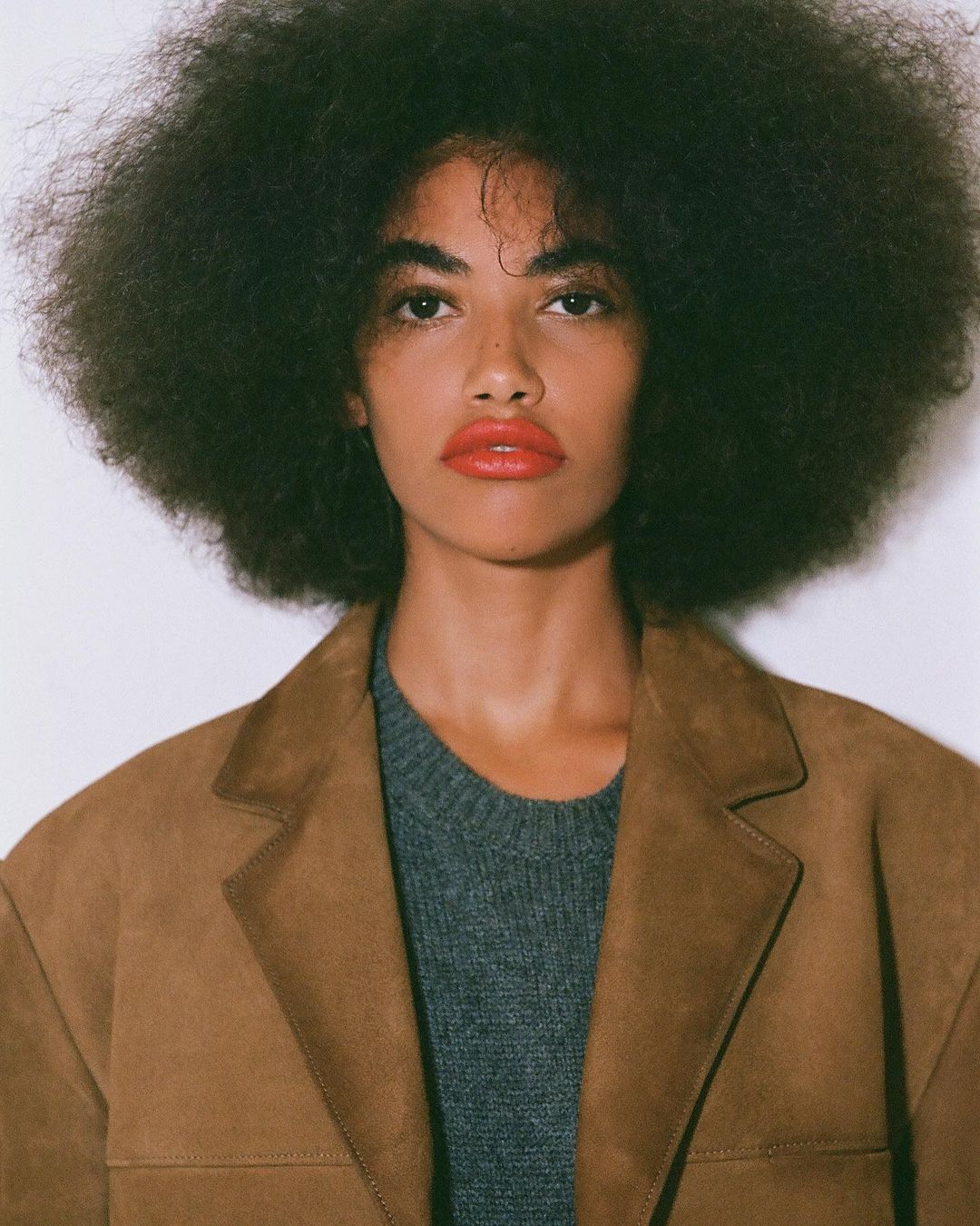 Portrait of a woman with a large afro hairstyle wearing a brown suede jacket over a grey sweater against a plain white wall