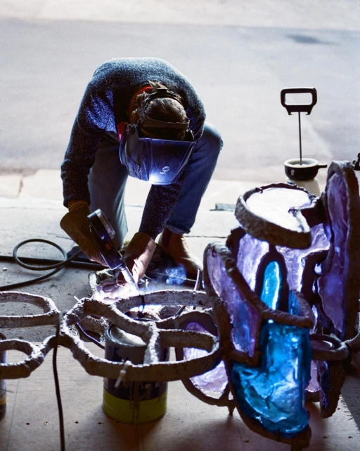 Adult working on a metal and glass art sculpture indoors, wearing a welding mask and gloves, using a welding machine. 