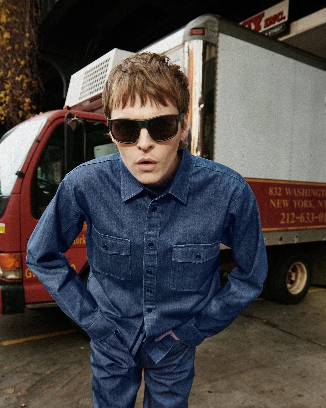 A person poses outside wearing sunglasses and a blue denim outfit, with a red and white delivery truck visible in the background.