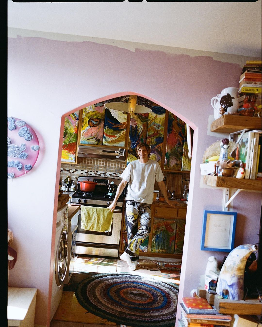 An adult stands in a kitchen filled with art, showcasing colorful paintings and a vibrant, artistic home interior with eclectic decor.