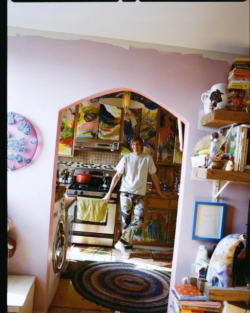 An adult stands in a kitchen filled with art, showcasing colorful paintings and a vibrant, artistic home interior with eclectic decor.