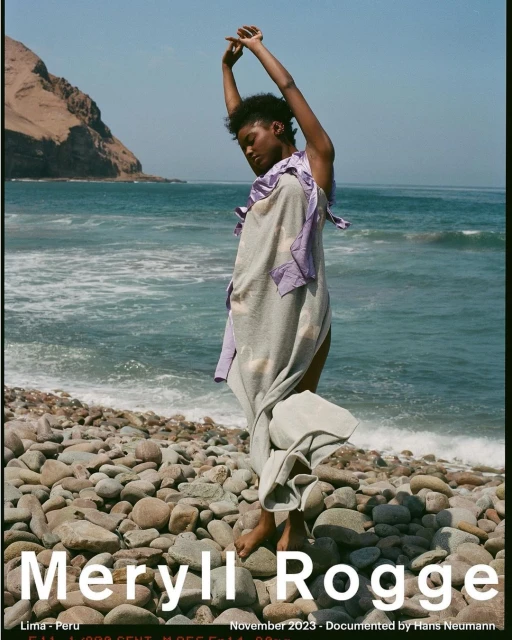 A barefoot fashion model poses on a rocky beach in a draped garment with ocean and mountains in the background on a sunny day.