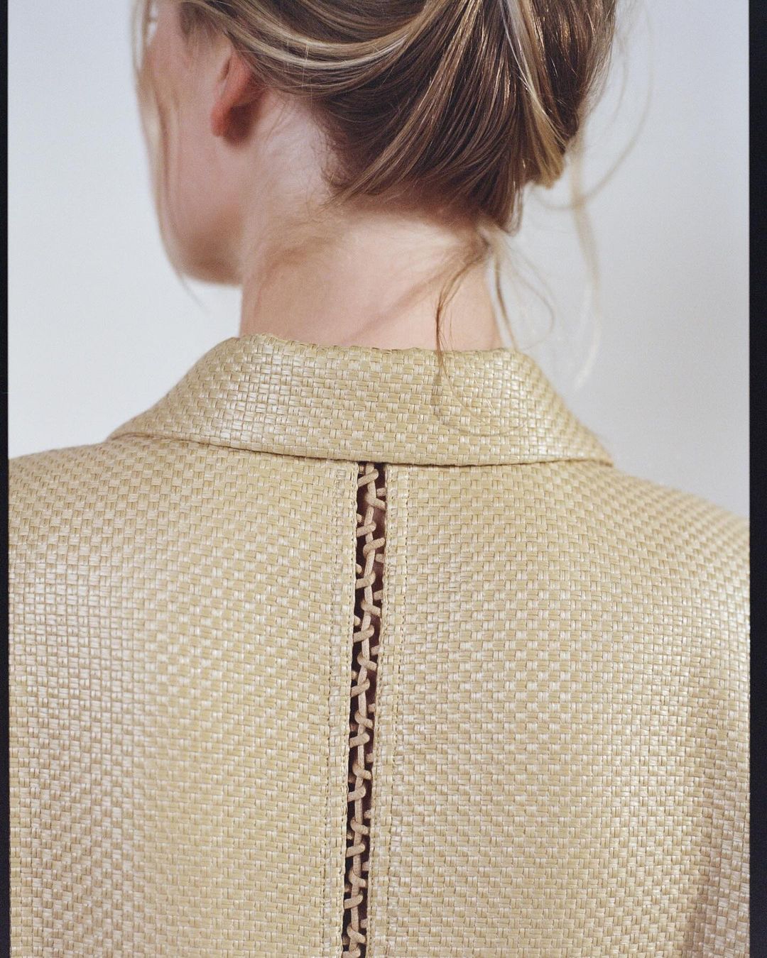 Woman with blonde hair in updo, back view of straw-textured jacket with detail, against a plain white background in portrait orientation.