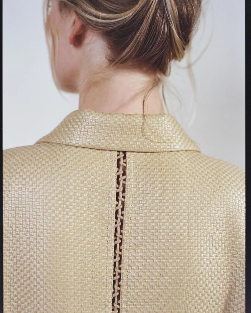 Woman with blonde hair in updo, back view of straw-textured jacket with detail, against a plain white background in portrait orientation.