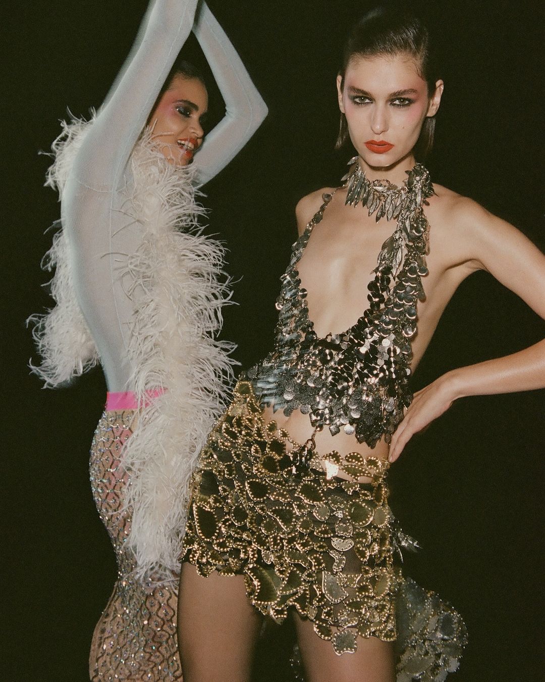 Two fashion models pose in a dark studio wearing shimmering, metallic dresses; one silver, the other gold, standing closely together.